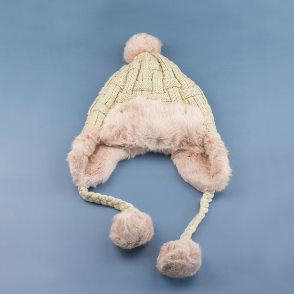 Beige cable-knit pom trapper hat laid flat on a blue background.