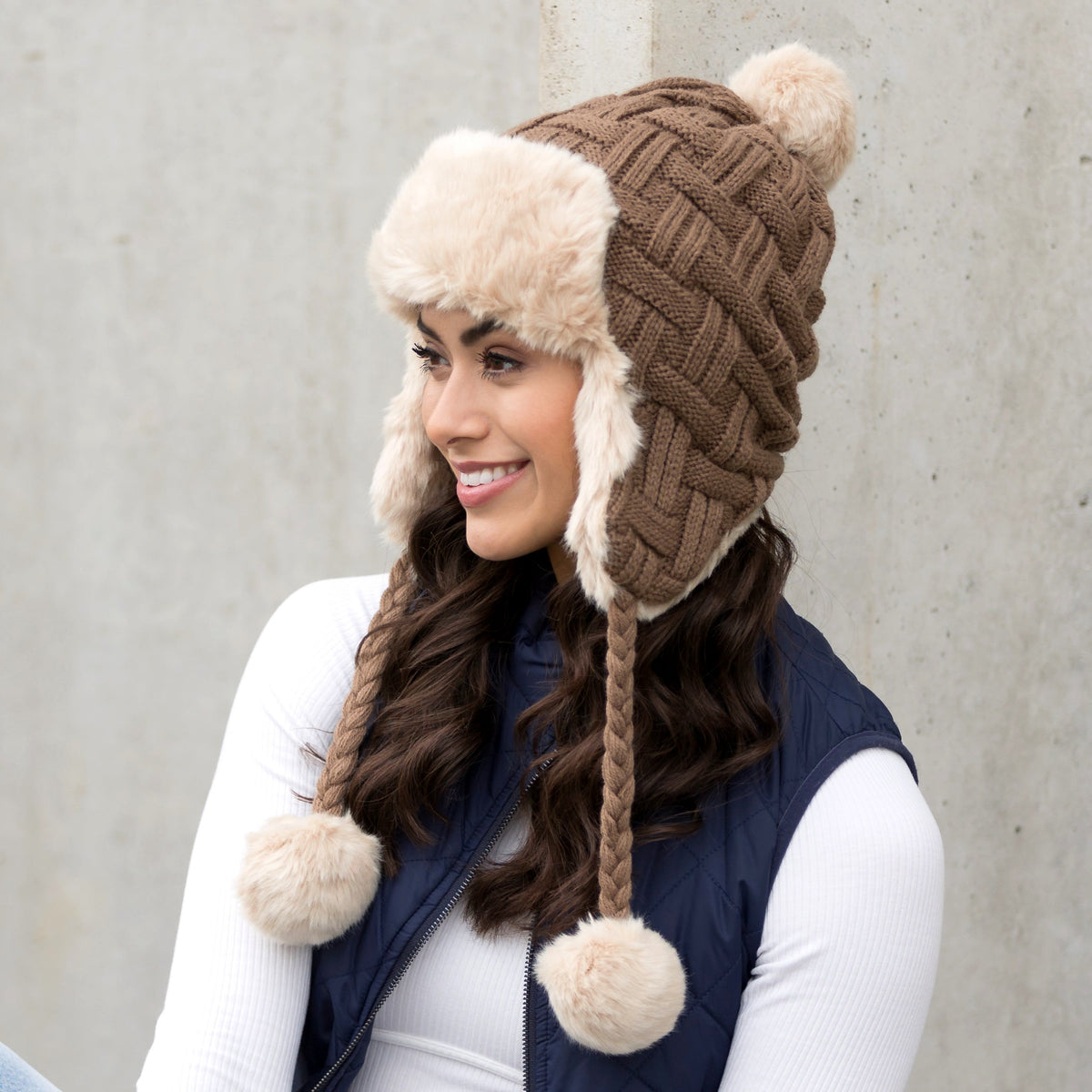 Taupe cable-knit trapper hat with light trim, ear flaps and dangling poms worn on a model.