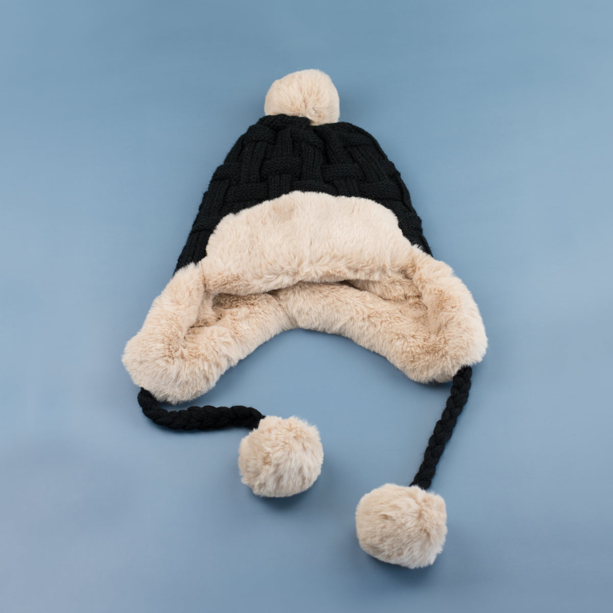 Black cable-knit pom trapper hat laid flat on a blue background.