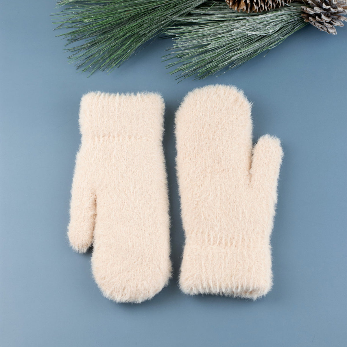 Almond-colored plush mittens laid flat beside evergreen sprigs.