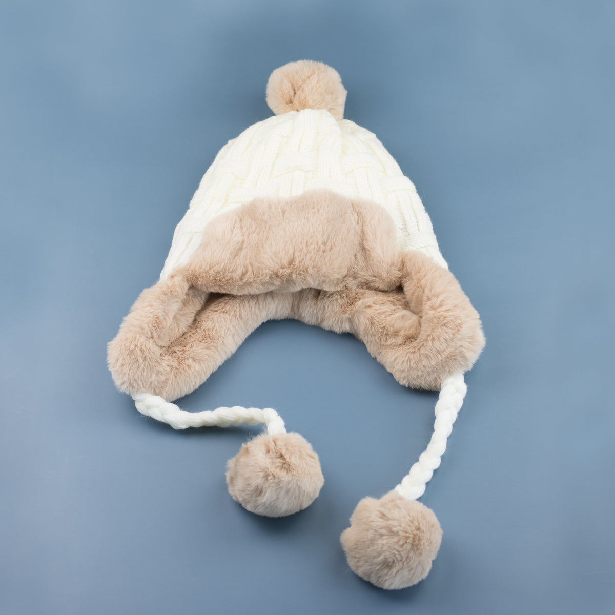Ivory cable-knit pom trapper hat laid flat on a blue background.