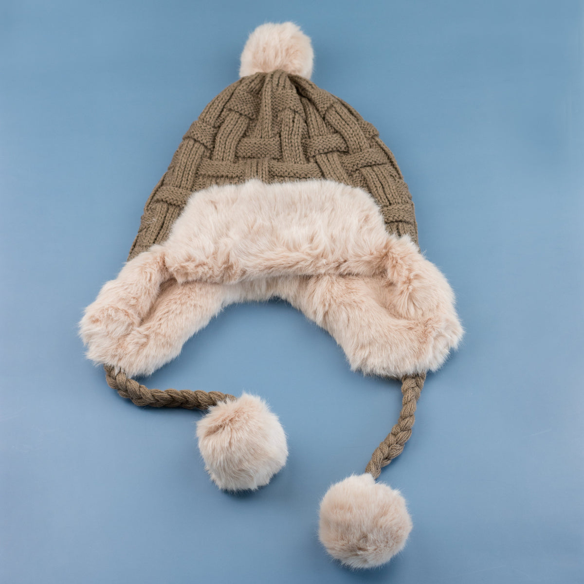 Taupe cable-knit pom trapper hat laid flat on a blue background.