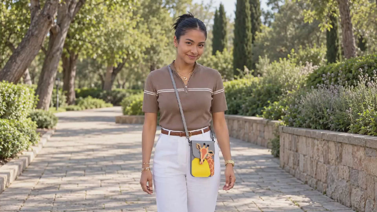 Woman wearing the gray and yellow Giraffe phone purse with its gray strap and black clips, gently touching the strap on a sunny park path. AI-assisted lifestyle scene.