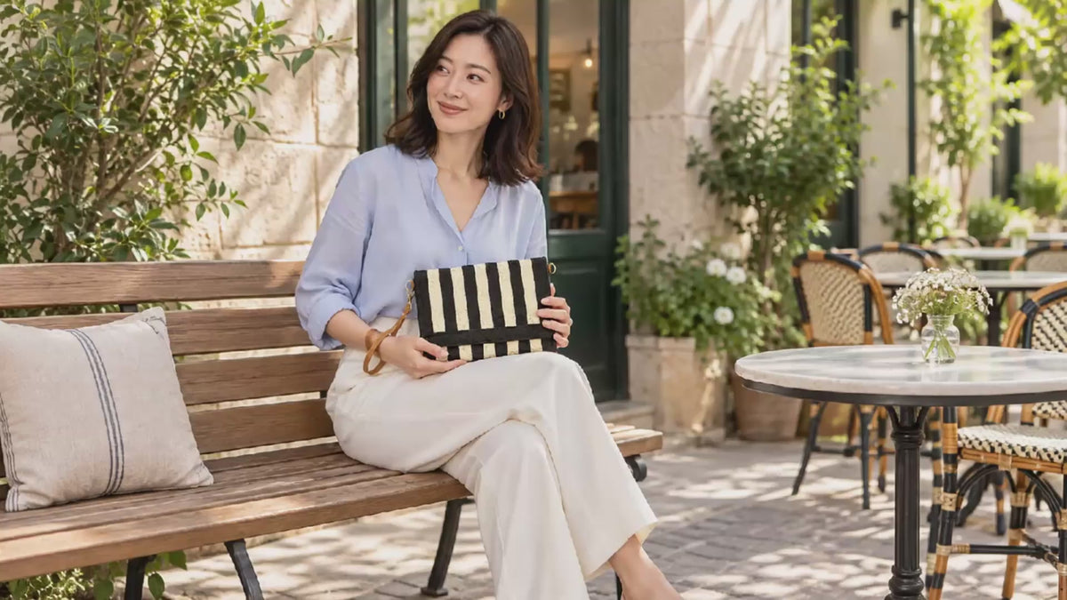 AI-generated lifestyle video of a model holding the black and cream Striped Woven Foldover Wristlet Crossbody Bag on a café bench.